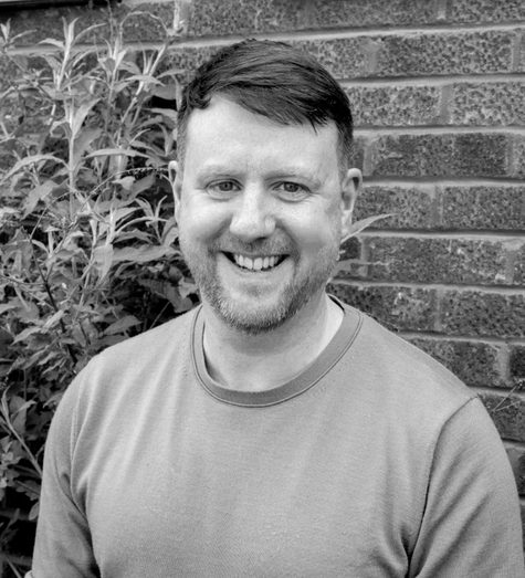 A black and white portrait of a smiling man with short hair and a beard, standing in front of a brick wall.