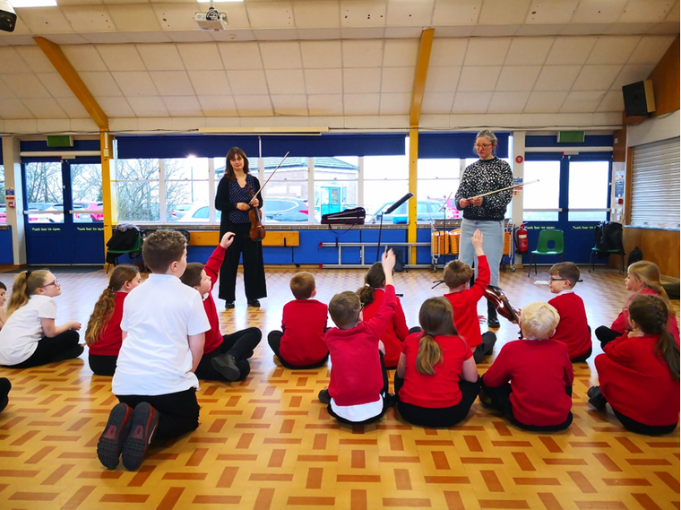A group of primary school children in red uniforms sit on a hall floor with their hands raised while two adult musicians stand at the front holding violins, leading an interactive music session.