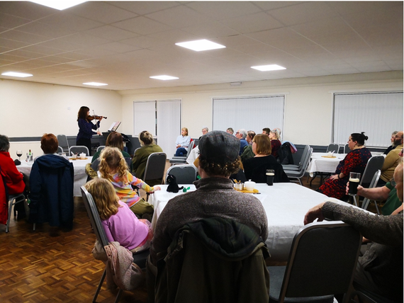 An adult violinist performs at the front of a community hall while adults and children sit around tables listening, some with drinks, in an informal concert setting.