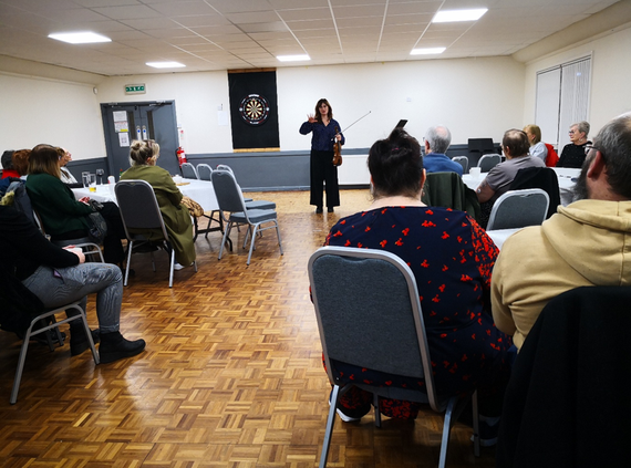 A violinist stands and speaks to a seated audience in a community hall, holding her instrument as people watch from chairs arranged around tables.