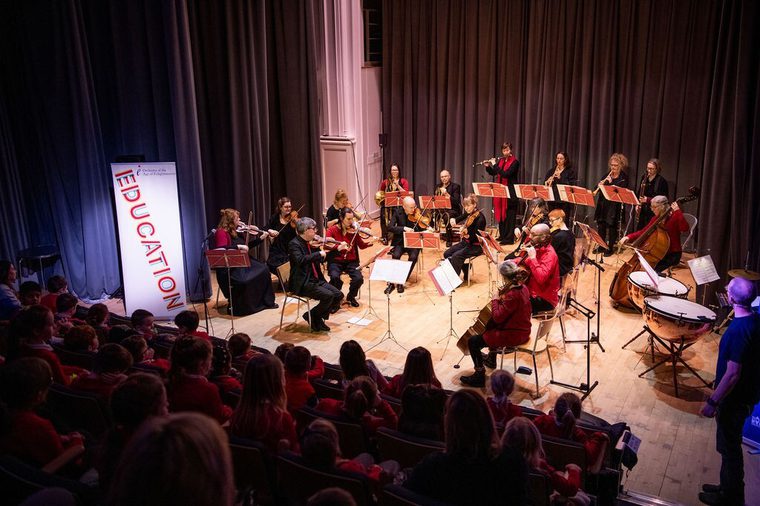An orchestra performs on a small stage before an audience of children. Musicians play string, wind, and percussion instruments, reading from sheet music.