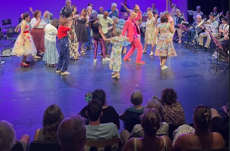 An intergenerational group of performers dancing on a stage in front of an audience, accompanied by live musicians in the background.