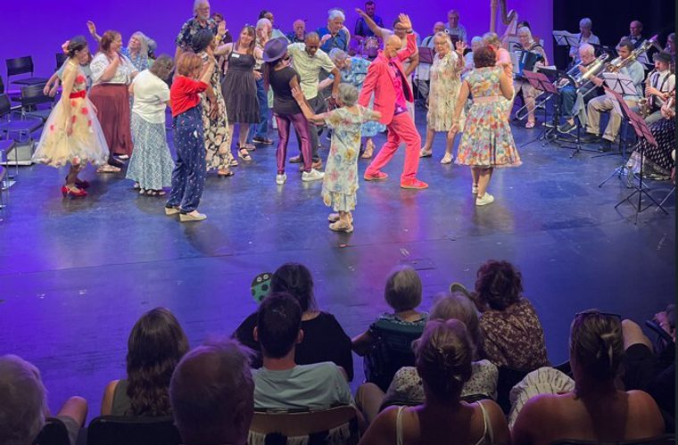 An intergenerational group of performers dancing on a stage in front of an audience, accompanied by live musicians in the background.
