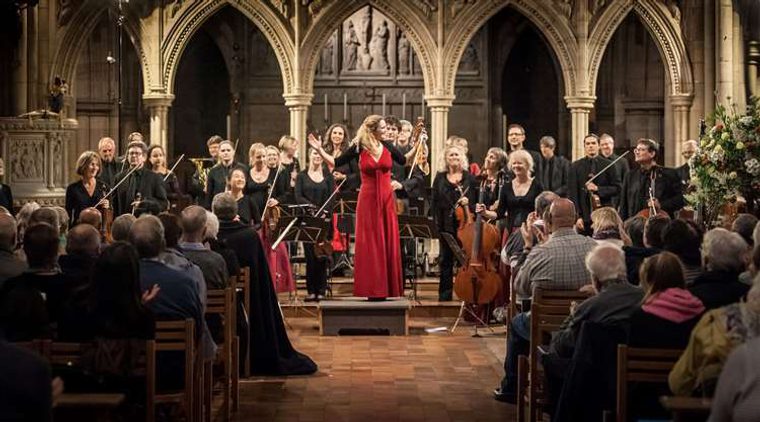 A chamber orchestra applauds a conductor in a red dress at the front of a church, while the seated audience claps, with stone arches rising behind the musicians.