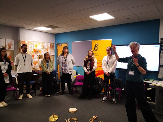 Group of adults standing in a workshop, led by an instructor gesturing while teaching a music activity.