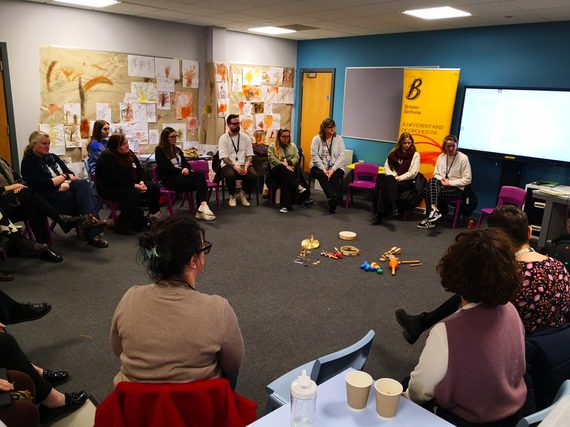 Adults seated in a circle in a classroom, discussing music with percussion instruments placed on the floor.
