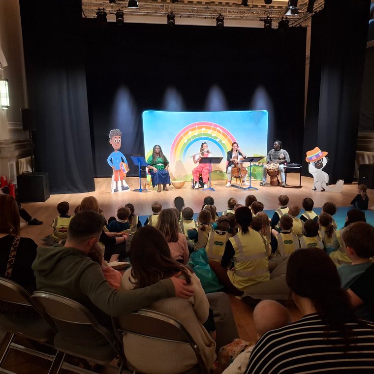 Musicians performing on stage for an audience of young children seated on the floor, with colourful backdrop.