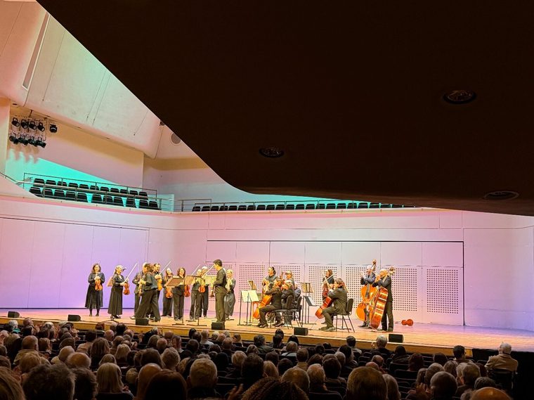 An orchestra performing on stage inside the Royal Concert Hall, with musicians playing string instruments in front of a seated audience.