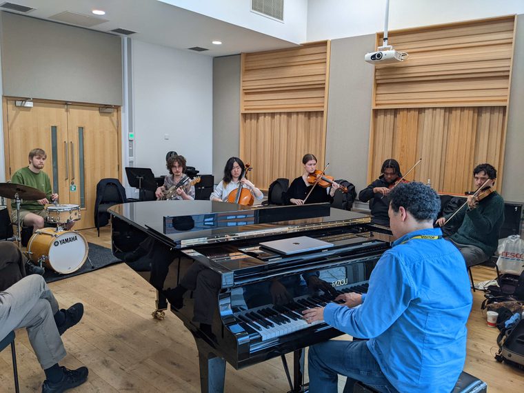 A group of young musicians rehearse together in a studio, playing piano, strings, bass and drums in a collaborative ensemble session.