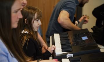 A girl sat at a keyboard in a room with other musicians,
