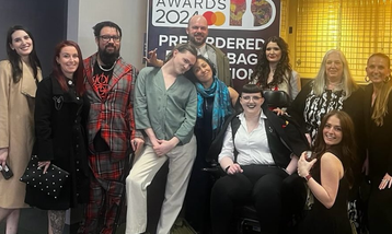 A group of people stand infront of a Brit Awards sign, smiling to the camera