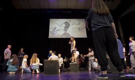 Performers on a stage, one woman in the centre stood on a box, a screen showing a video at the back of the stage