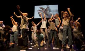 A group of performers on stage with arms outstretched in different directions. A screen behind showing a hand reaching out.