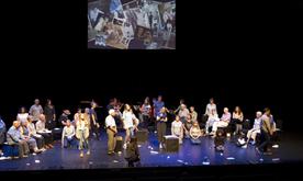 A group of performers on stage with a screen behind showing a film