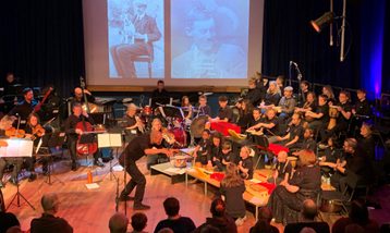 Orchestra of professional musicians and young musicians performing on stage with screen projections behind them