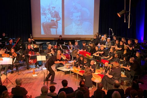 An orchestra made up of professional musicians and young musicians performing on stage with projections on a screen behind them