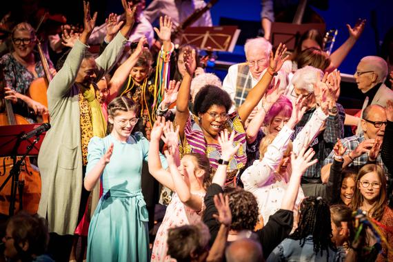 Group of people on stage with musicians. They are all happy and have their arms in the air
