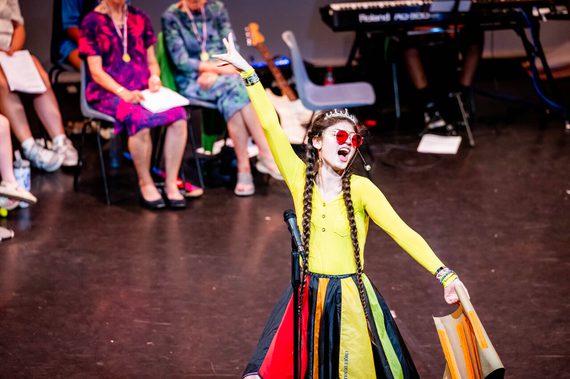 A young woman in a bright dress sings confidently with one arm raised