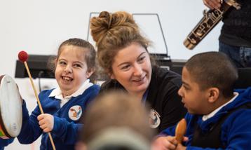 Two young children playing percussion instruments with a school teacher.