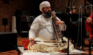 Image of a man of Asian heritage sitting on a stage with an Indian musical instrument in his left hand and a bow in his right hand