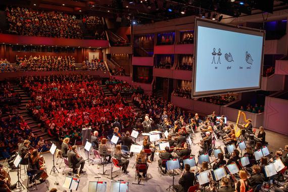 A full orchestra on stage performing to an audience of hundreds of school children, with a big screen overhead.