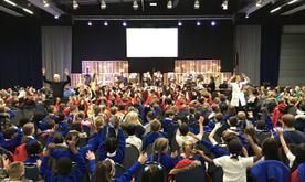 A large audience of young children waving at two presenters stood in the aisles of the venue.