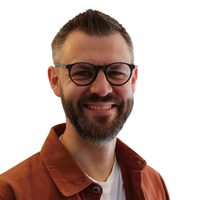 Headshot of Giles Wright, Chair of Orchestras Live, wearing a white t-shirt and orange over shirt. He is white and has a beard, glasses and short brown hair