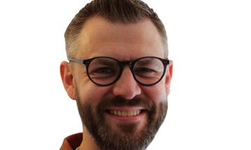 Headshot of Giles Wright, Chair of Orchestras Live, wearing a white t-shirt and orange over shirt. He is white and has a beard, glasses and short brown hair