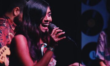 A woman on stage sings into a microphone with a joyful expression. She has long dark hair and is wearing a colorful outfit and a wristband. A guitarist and keyboard player are partially visible beside her under dim stage lighting.