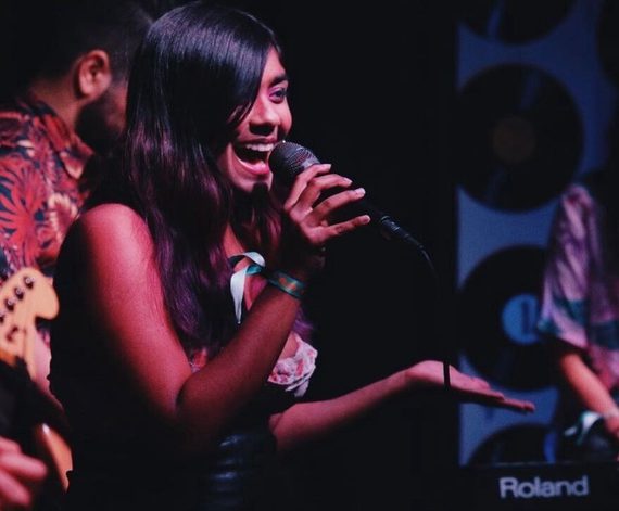 A woman on stage sings into a microphone with a joyful expression. She has long dark hair and is wearing a colorful outfit and a wristband. A guitarist and keyboard player are partially visible beside her under dim stage lighting.