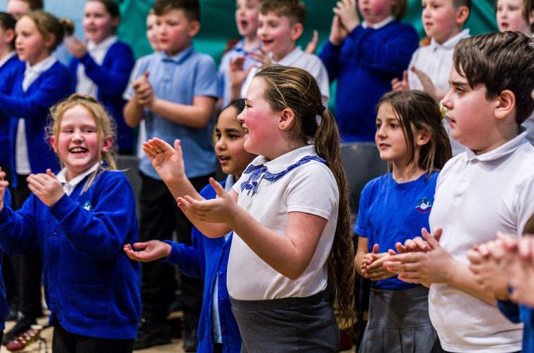 Primary school children in blue uniforms smiling and clapping during a performance.