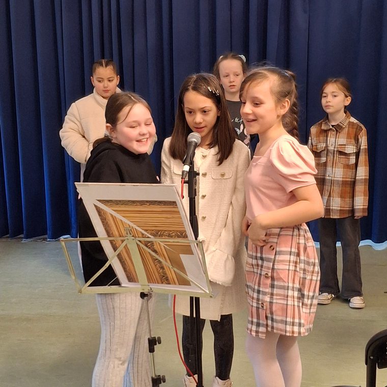 Three children stand at a microphone reading from a music stand, performing in front of a blue curtain.