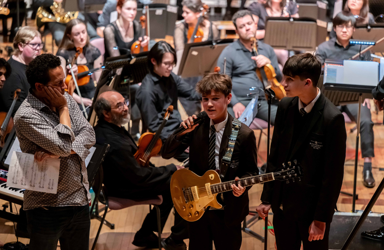 A student holding an electric guitar speaks into a microphone, standing between a classmate and an orchestra member during a stage performance.