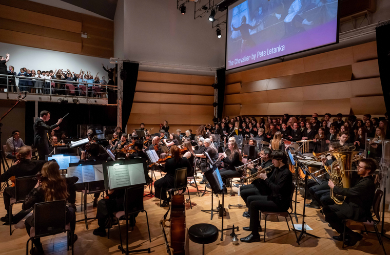 A large orchestra and choir perform in a concert hall below a screen displaying "The Chevalier by Pete Letanka."