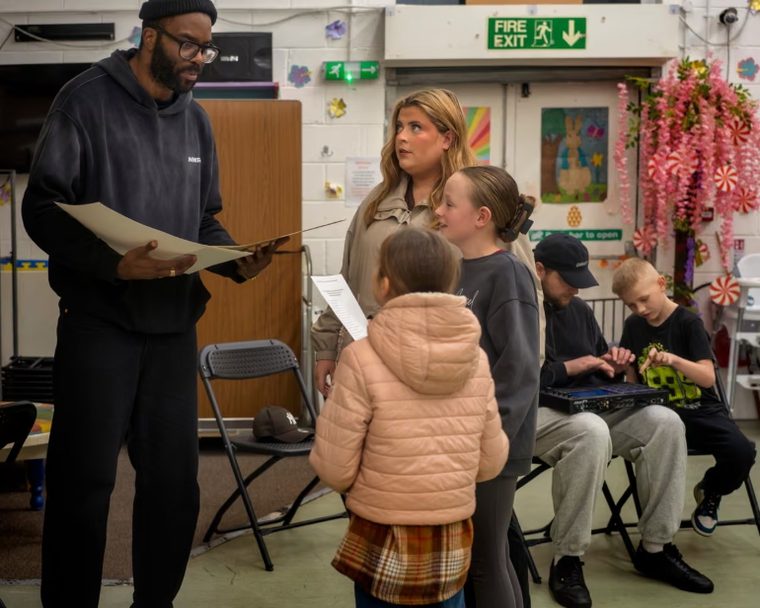An instructor holds a large sheet of paper while speaking to a woman and two young girls in an indoor workshop setting, as two other individuals sit in the background.
