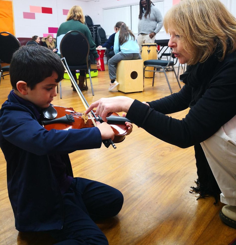 Music teacher guiding a young boy as he learns to hold and play a violin in a classroom setting.