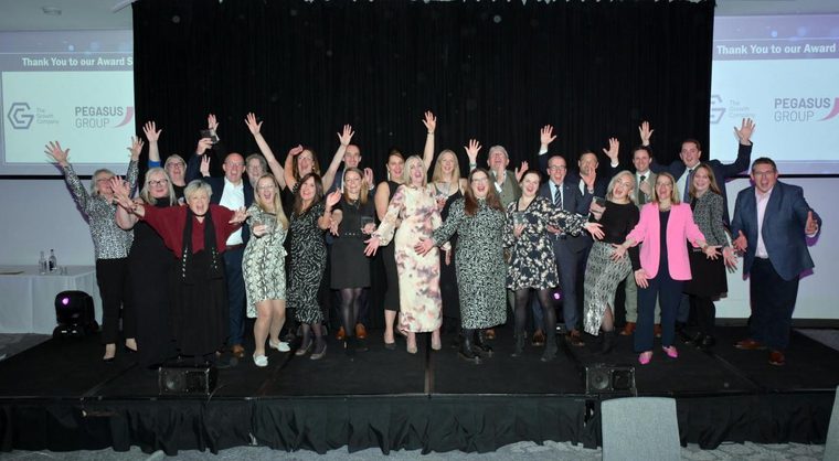 A large group of people from East Riding of Yorkshire Council standing together on a stage at an awards event, smiling and raising their arms in celebration. The group is dressed in formal and semi-formal attire. Behind them, screens display event sponsor logos including ‘The Growth Company’ and ‘Pegasus Group.’ The atmosphere is lively and celebratory.